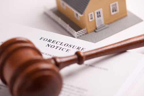 A small peach colored house in the background on a white table. A foreclosure notice lays on the table with a wooden gavel on top for Foreclosure